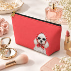 Red makeup pouch with a white Poodle in sunglasses and a red scarf beside perfume and makeup