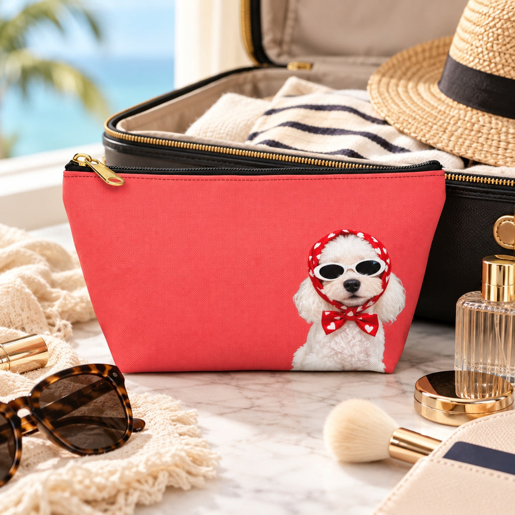 Red makeup pouch with a white Poodle in sunglasses and a red scarf beside a suitcase and straw hat