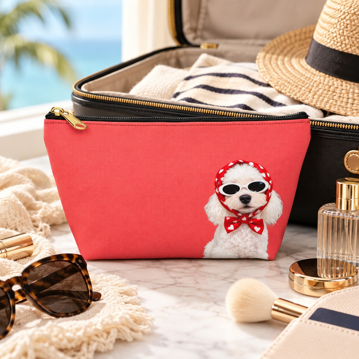 Red makeup pouch with a white Poodle in sunglasses and a red scarf beside a suitcase and straw hat